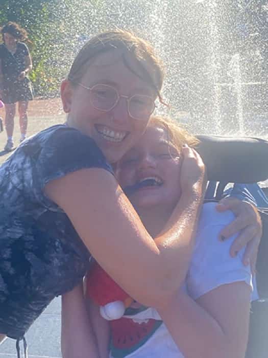 Fachkraft umarmt ihr lachendes Gastkind im Rollstuhl vor einer Wasserfontäne